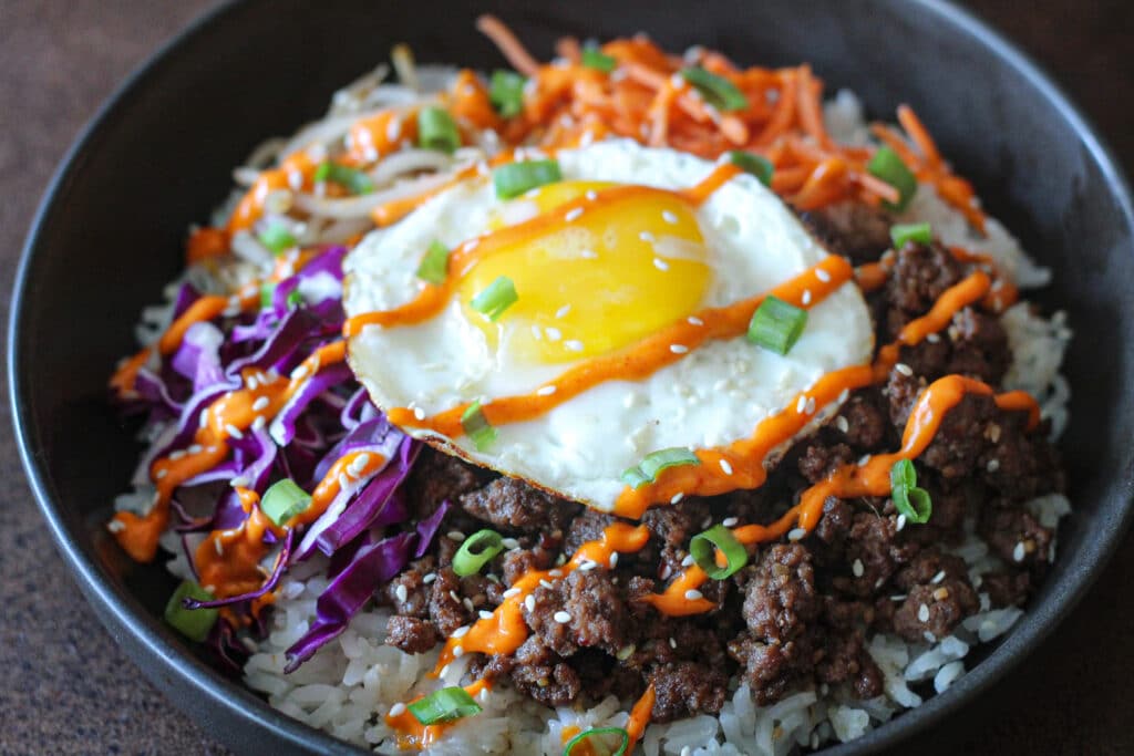 Korean ground beef bulgogi bibimbap with jasmine rice, ground beef, fried egg, red cabbage, bean sprouts, shredded carrots, green onion, sesame seeds, and a spicy gochujang sauce